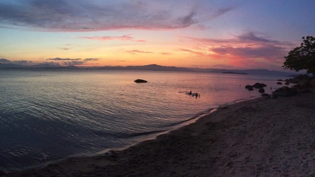 An evening on a sand beach with the last tones of pink and yellow left by a sunset on the darkening sky. Two people are swimming in the ocean close to the beach line with some rocks situated on the back.