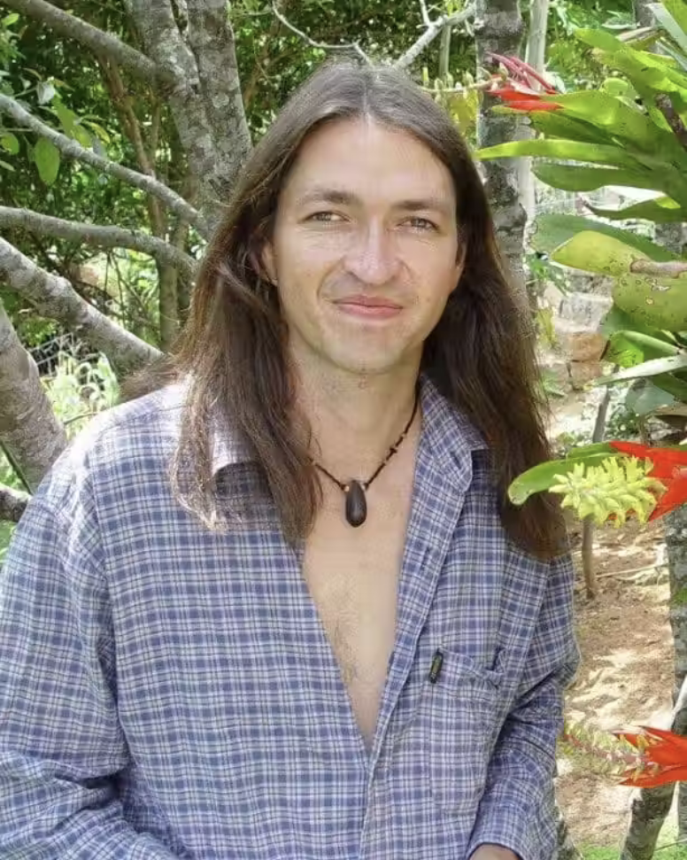 A man with long hair standing in front of a tree.