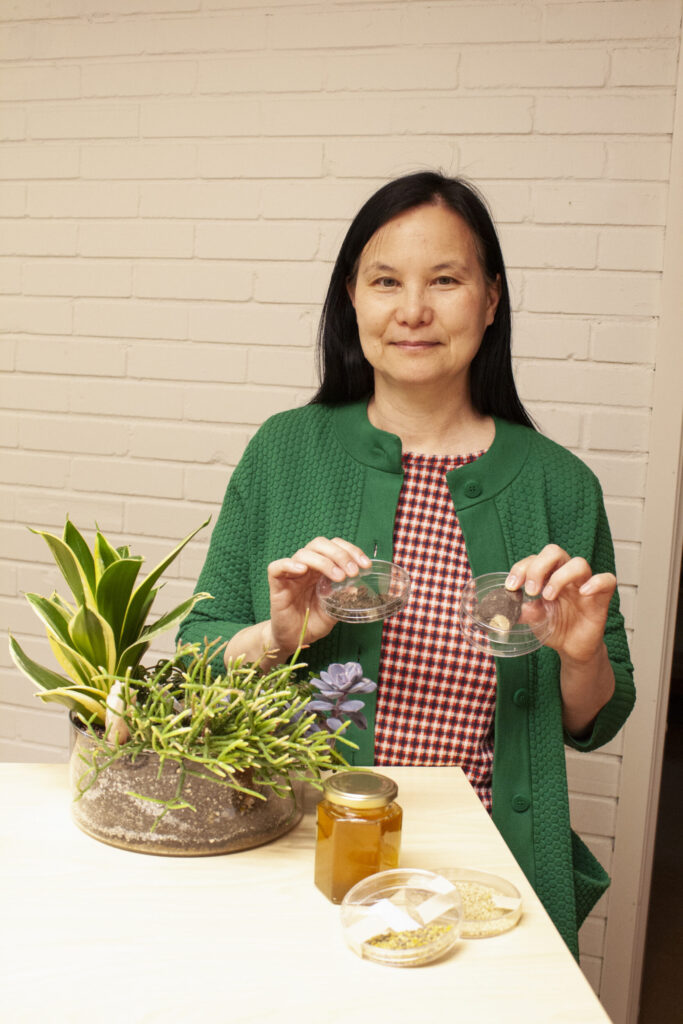 A woman in a green cardigan and plaid shirt sitting at a table, smiling at the camera while holding two small jars of seeds with a potted plant arrangement beside her.