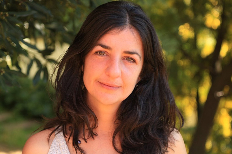 A woman with dark hair and a gentle smile looks directly at the camera, set against a blurred greenery background. she has a subtle, warm expression.