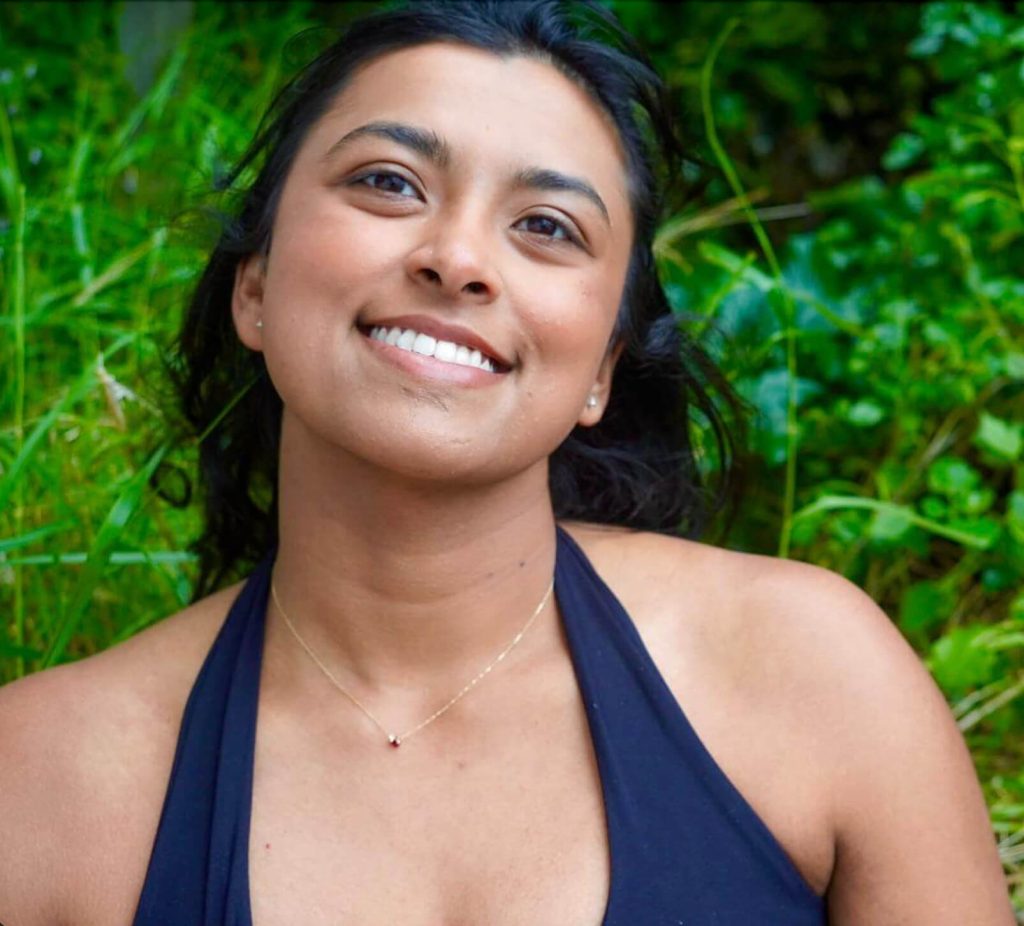 A woman with dark hair and a black halter top smiles outdoors, surrounded by lush green foliage.