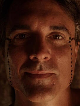 A person with short hair looks directly at the camera, illuminated by warm light. There are black dotted lines drawn vertically from their eyes down to their jawline on both sides of their face.
