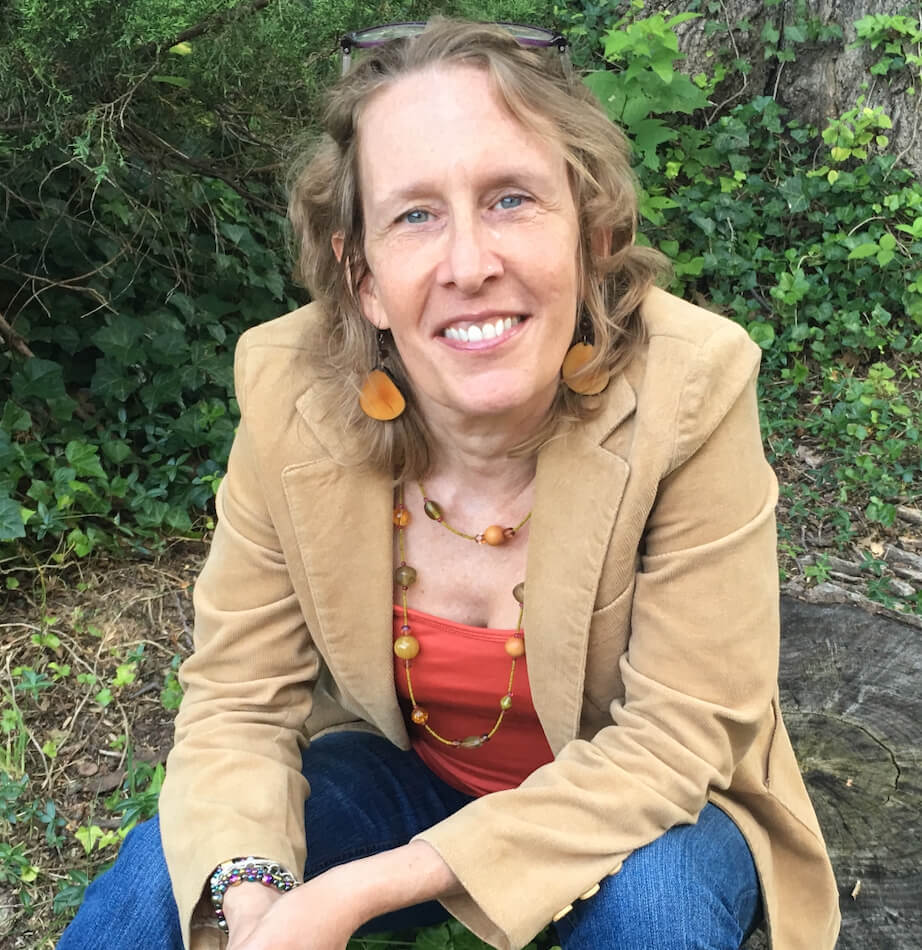A woman with wavy blonde hair, wearing a tan blazer, orange top, blue jeans, and amber jewelry, sits outdoors on a rock, smiling at the camera with greenery in the background.