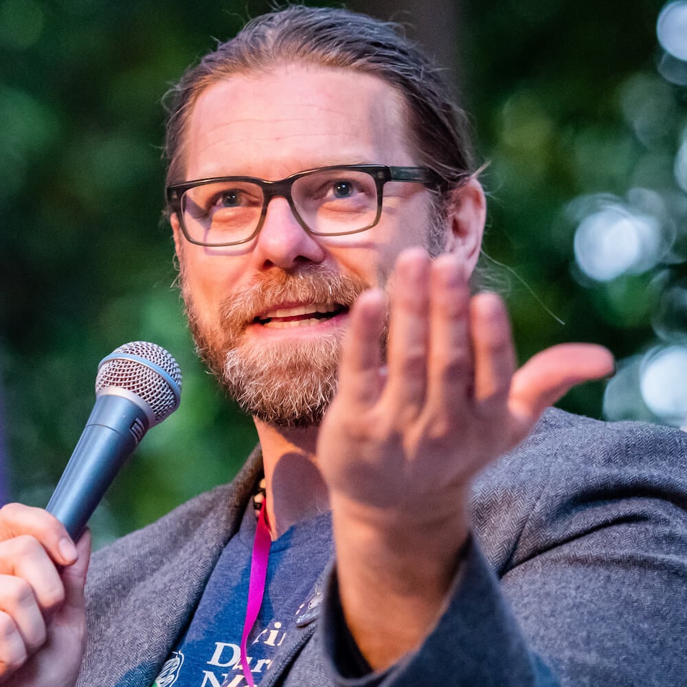 A bearded man with glasses holds a microphone and gestures with his other hand while speaking outdoors, with green foliage blurred in the background.