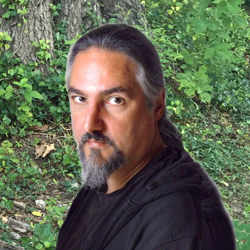 A man with long dark hair and a gray beard is wearing a black shirt and looking at the camera. He stands outdoors in front of green foliage and a tree trunk.